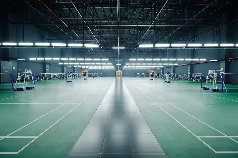 An empty indoor sports hall with badminton courts marked on a green floor under rows of lights.