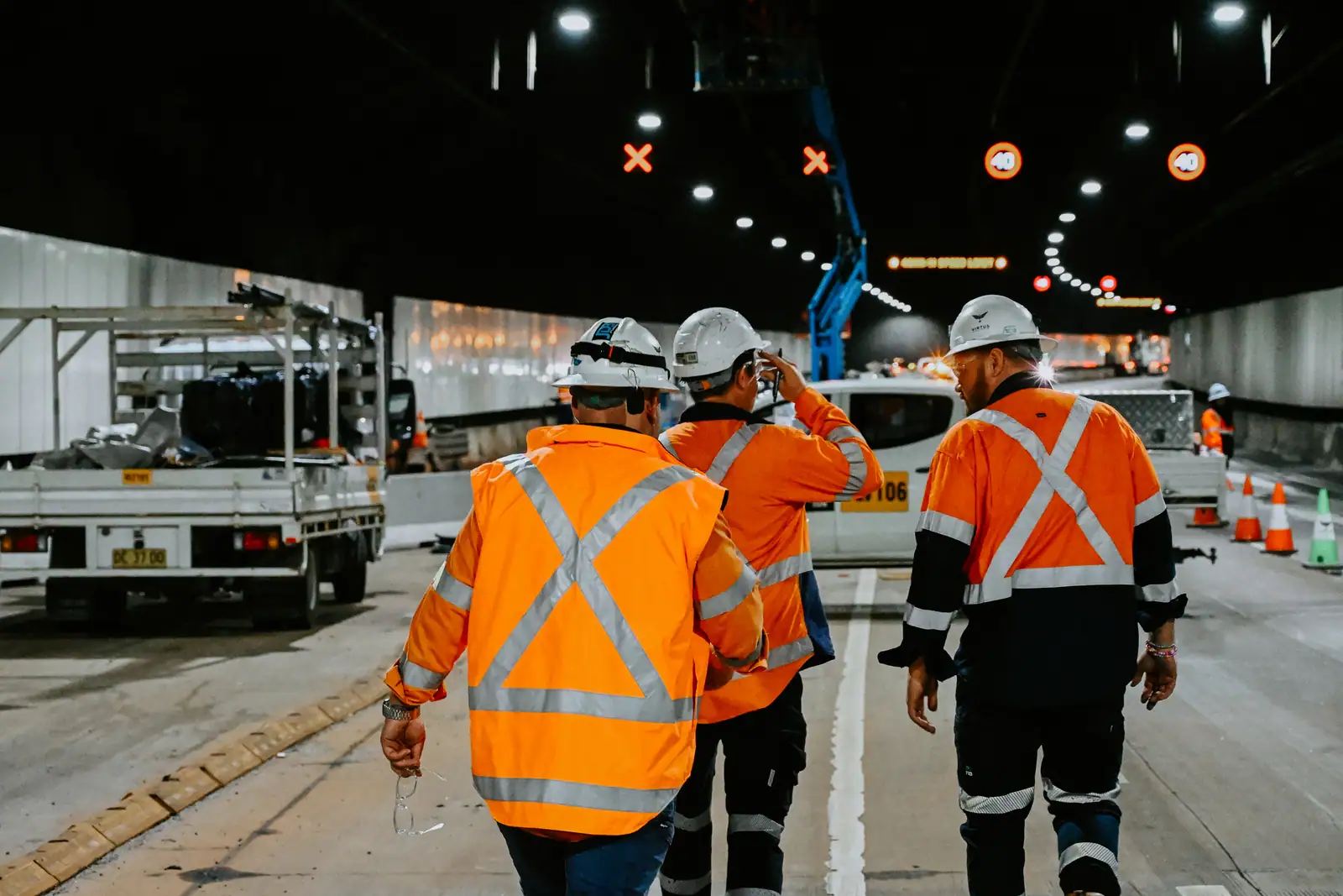 Three Virtus crew in high-visibility vests walking down the closed tunnel carriageway under the lane signals.