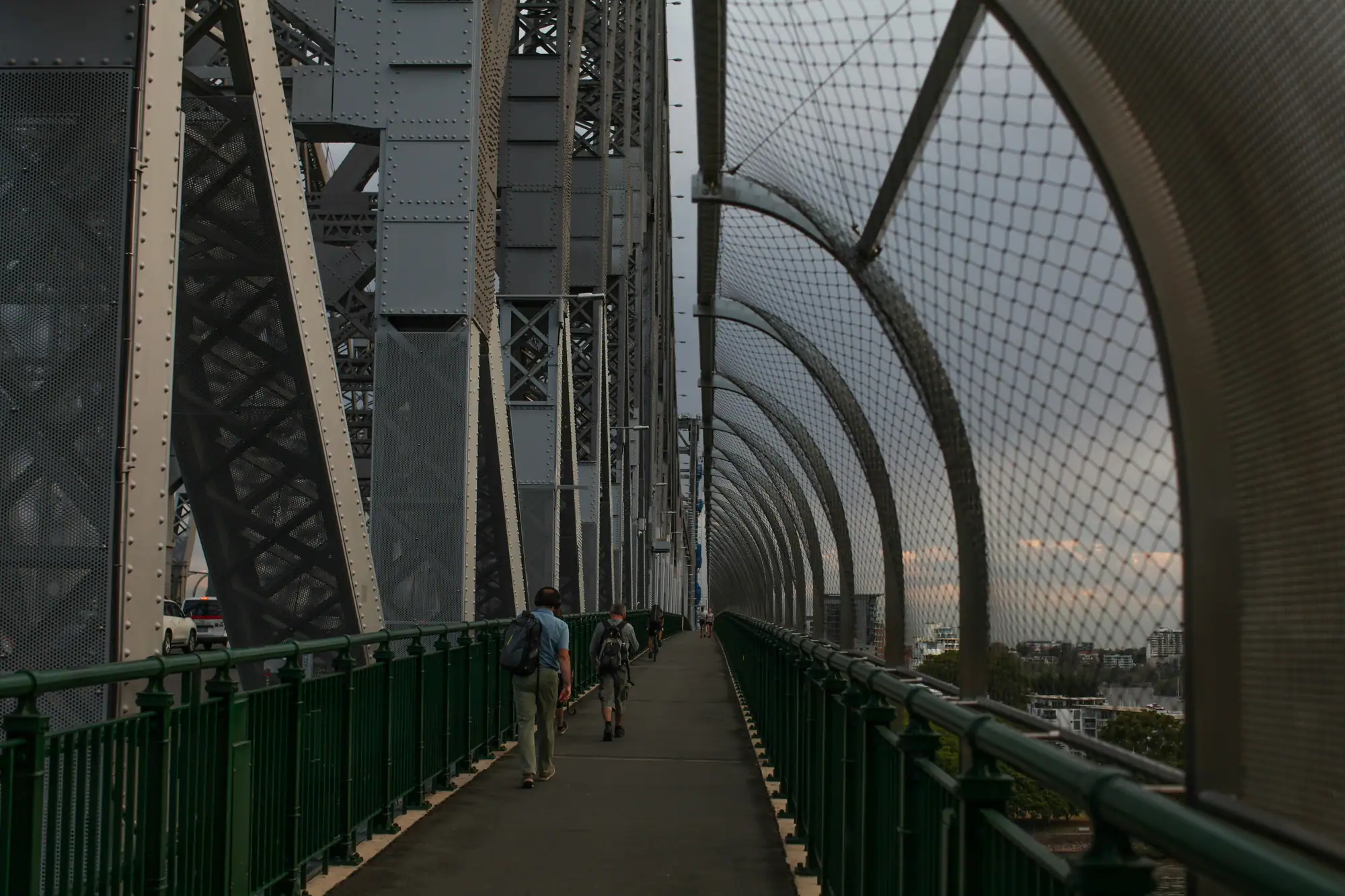 Pedestrians on the walkway of a riveted steel truss bridge at dusk, safety mesh arching overhead