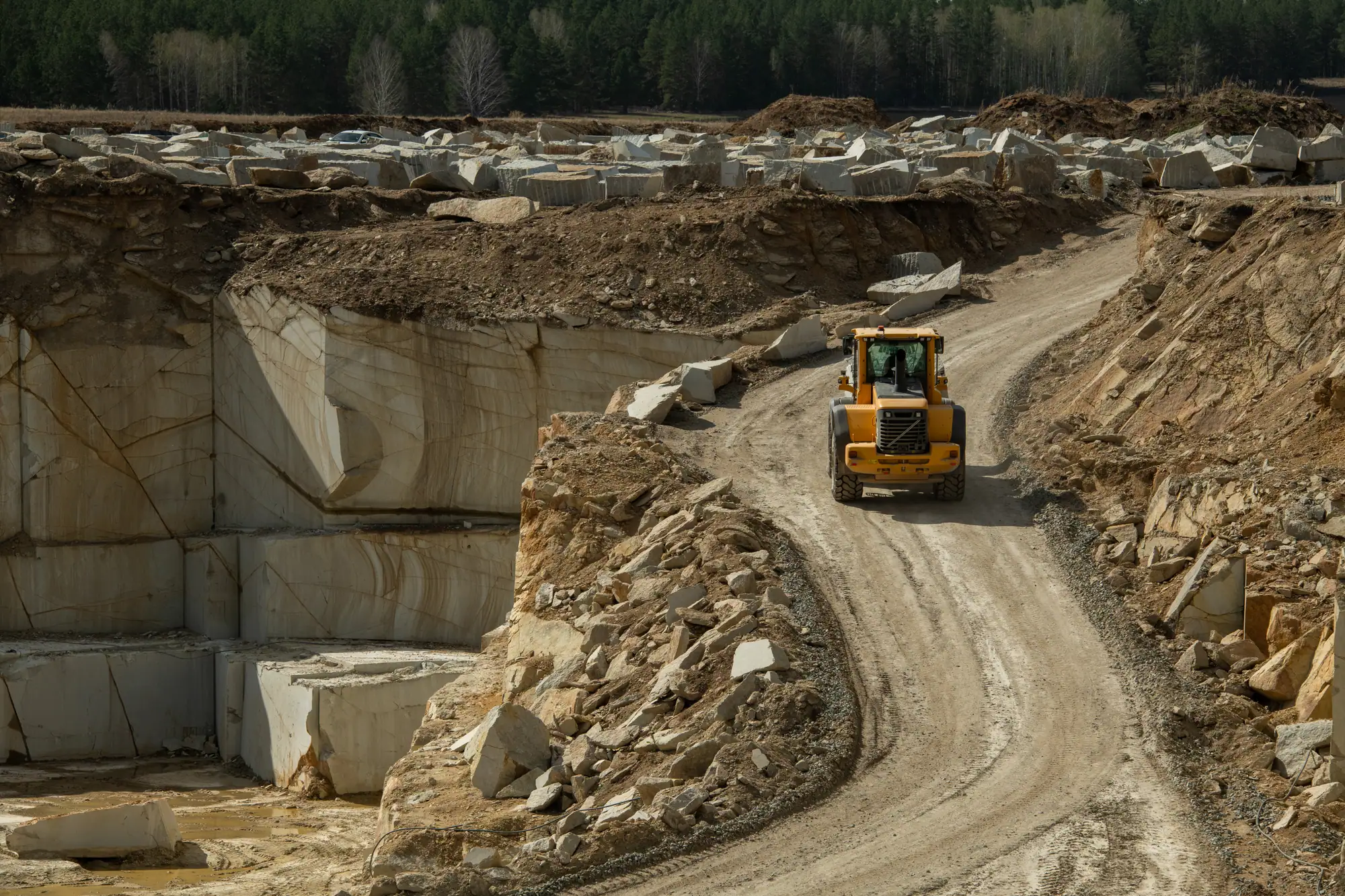 Yellow loader on a haul road winding through an open-cut quarry with cut stone blocks stacked above