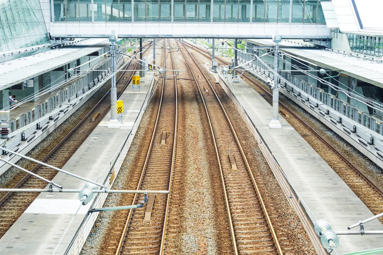 Railway corridor seen from above the tracks: three lines converge towards a station under overhead catenary, with platforms and canopies either side.