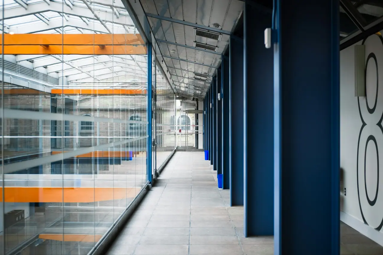 Glazed walkway inside a commercial building: steel roof structure above, aluminium-framed glass partitions to the left and a row of blue-framed doors to the right.